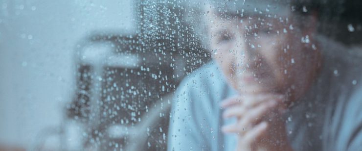 אישה מבוגרת משלבת את כפות ידיה ובוהה בקיר זכוכית רטוב